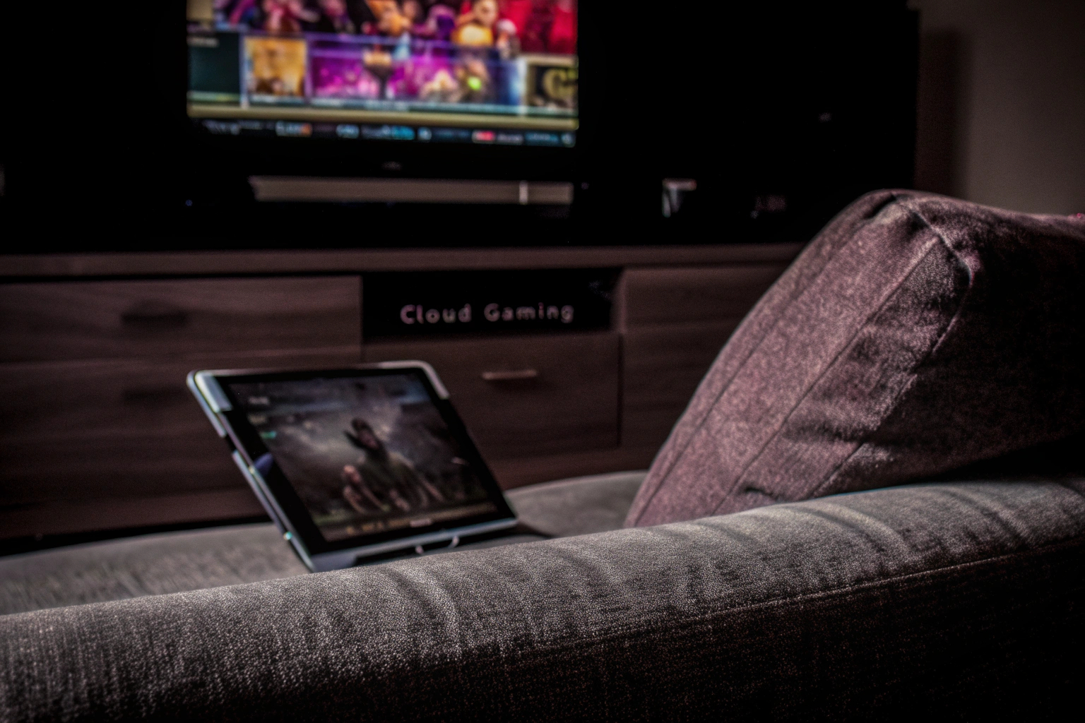 Ein Gamer, der auf einem Tablet Cloud Gaming spielt, während er auf einem Sofa sitzt. Im Hintergrund ist ein Fernseher, der ebenfalls ein Spiel zeigt. Gamer spielt Cloud Gaming auf einem Tablet